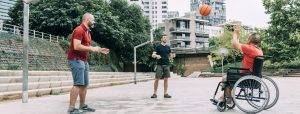 Image of three men at a basketball court playing basketball. One man is in a wheelchair and the other two are standing. They are all smiling.